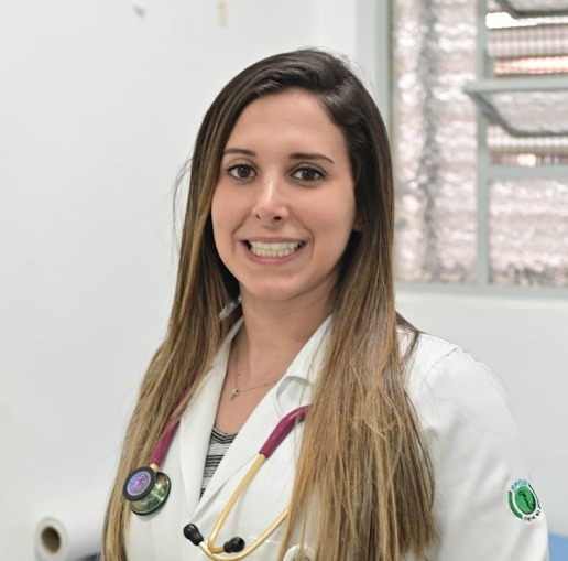Na imagem, a médica Natália Gonçalves tem cabelos longos, está um jaleco branco e com um estetoscópio em volta do pescoço. Ela está sorrindo para a foto.